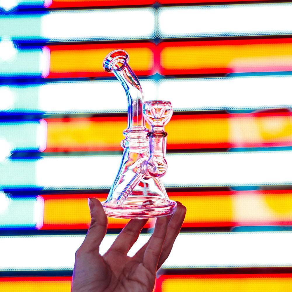 Hemper x Lil Debbie Rig held in hand, clear glass with 14mm joint, against colorful striped background