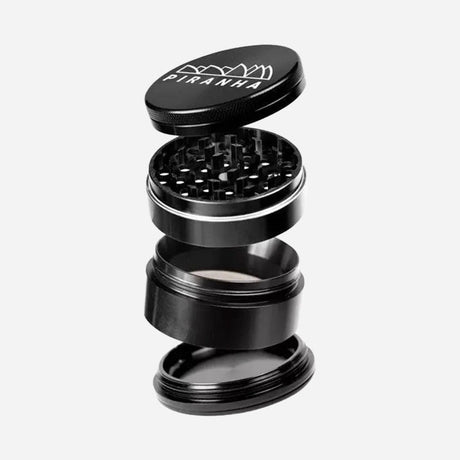 A black 4-part herb grinder with a sleek metal design. The top lid, engraved with "Piranha" and a shark-tooth logo, opens to reveal sharp grinding teeth. Below are a mesh screen for pollen collection and a storage chamber.