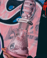 16" AFM Glass Beaker Bong with Freezable Coil and Reversal UFO Pyramid, held against a graffiti background