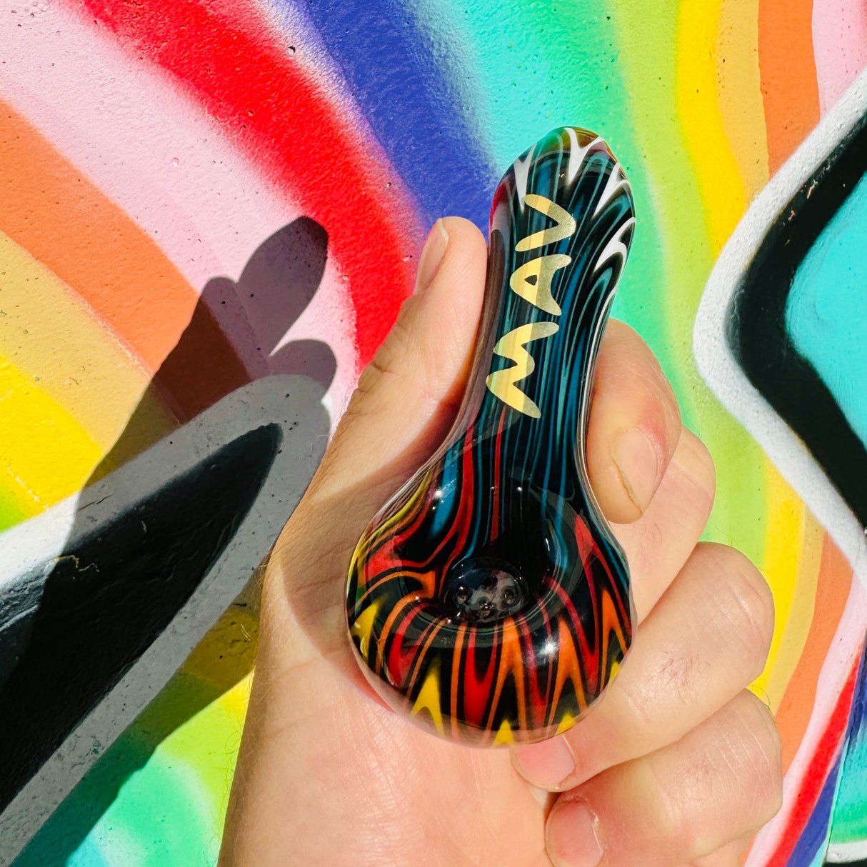 MAV Glass wigwag golden Professional Hand Pipe with vibrant color swirls, held in front of a graffiti wall