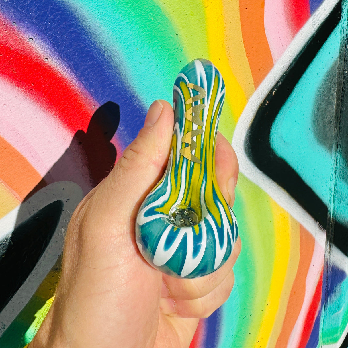 MAV Glass wigwag golden Professional Hand Pipe with colorful swirl design, held in front of graffiti wall