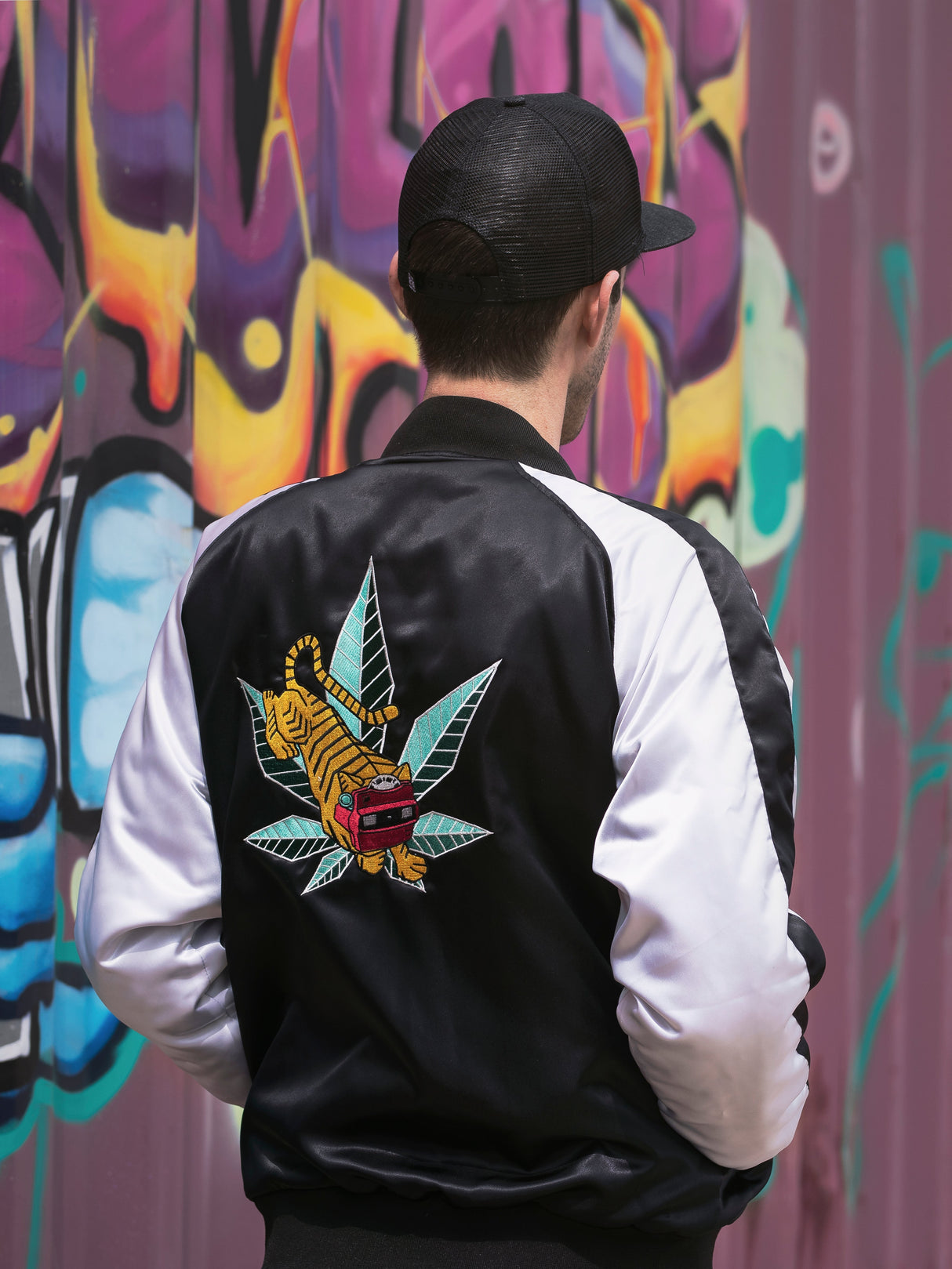 Person wearing GRAV Satin Bomber Jacket with logo on back, standing in front of graffiti wall